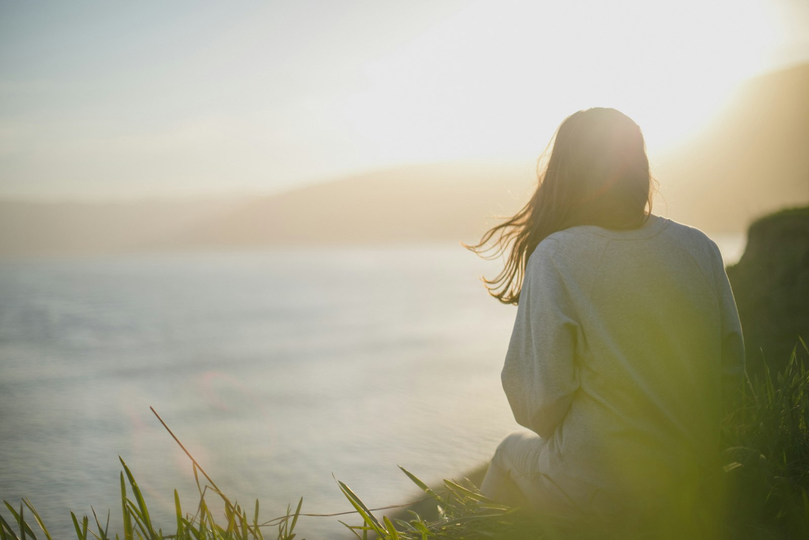 Une femme de dos, assise dans l'herbe face à la mer, cheveux au vent, dans la lumière du matin