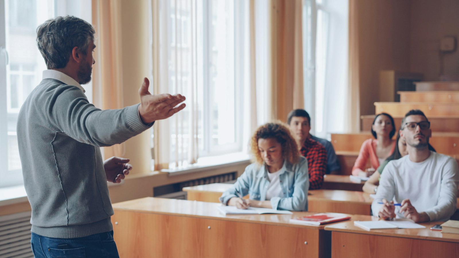 Un enseignant debout, le bras ouvert, s'adresse à des élèves assis à leurs pupitres dans une salle lumineuse