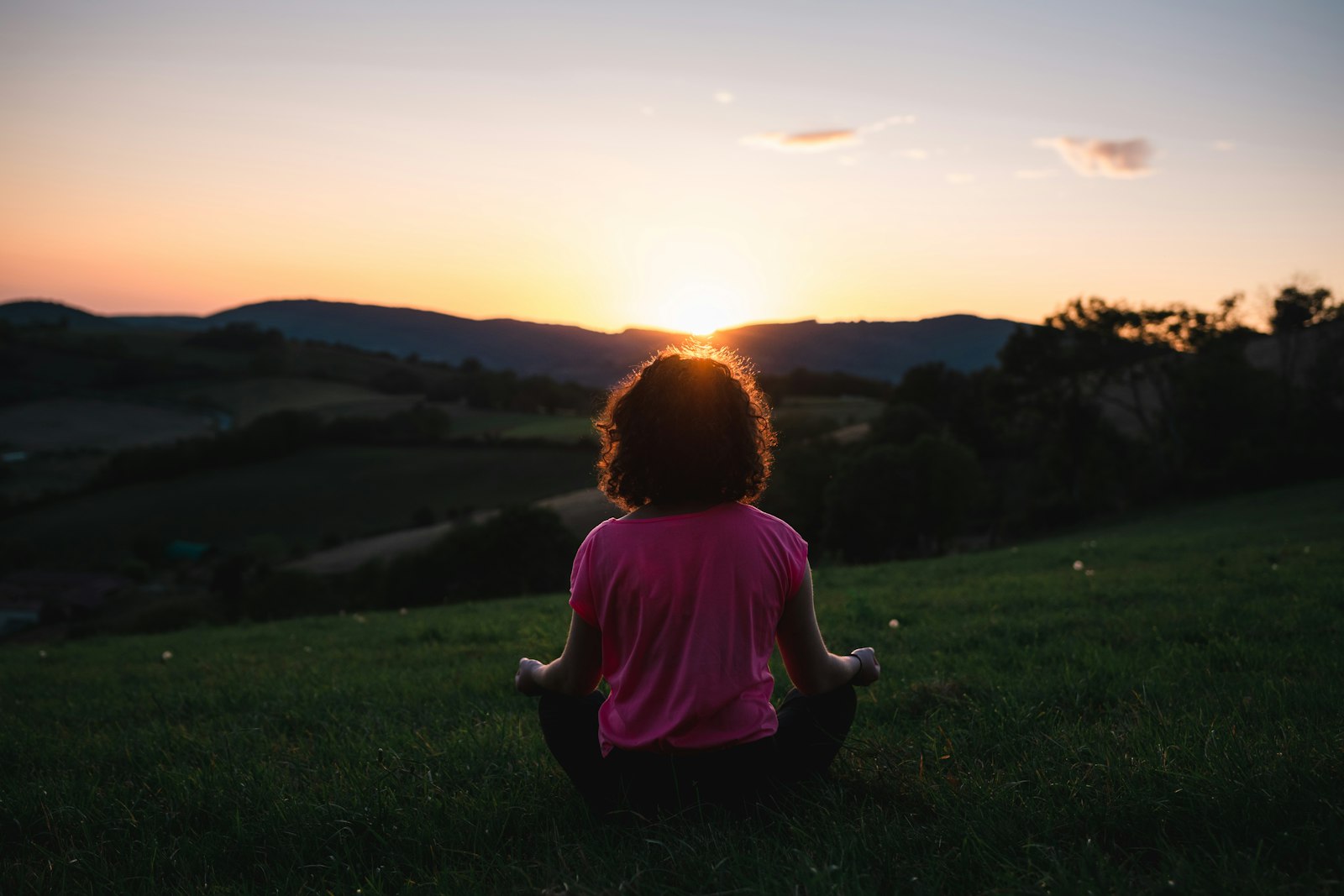 Une personne assise en tailleur dans l'herbe, de dos, face à un coucher de soleil sur des collines