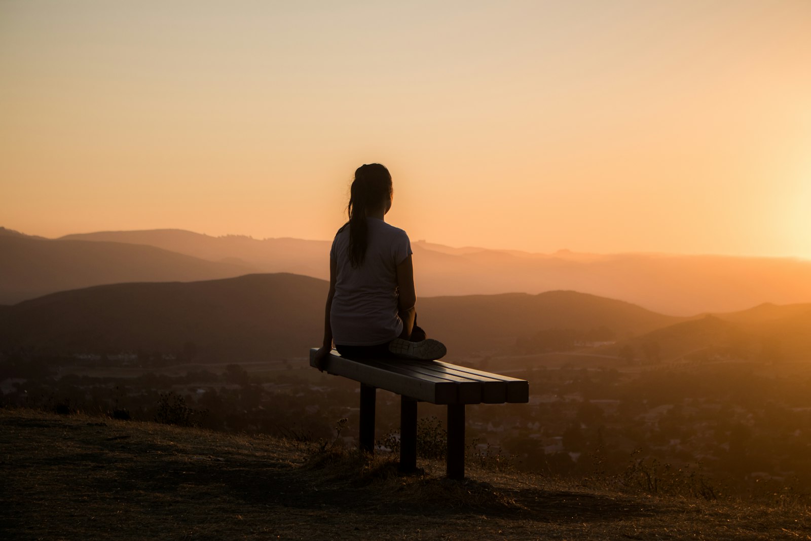 Une femme assise sur un banc regarde des collines au lever du soleil, vue de dos