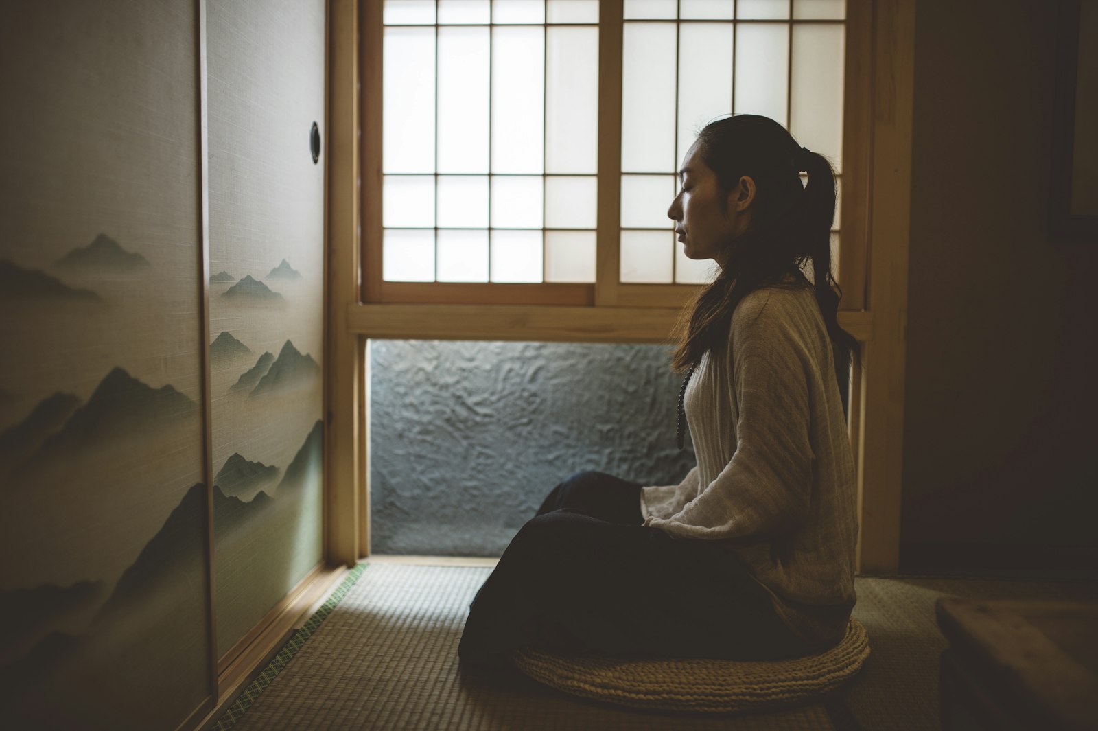 Une femme assise en silence près d'une fenêtre, les yeux fermés, dans un moment de calme et de concentration