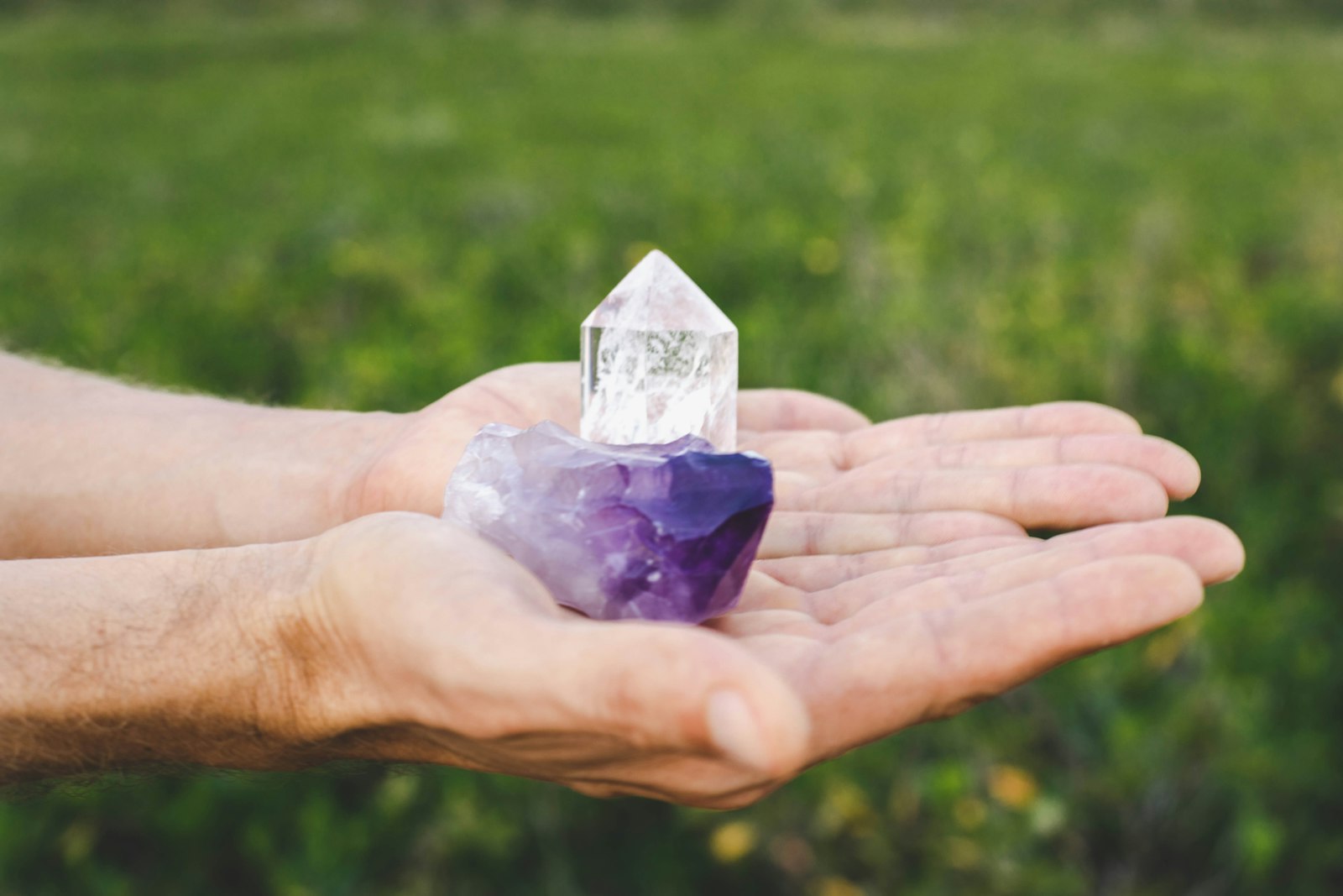 Deux mains ouvertes présentent un morceau d'améthyste surmonté d'un cristal de roche, sur un fond d'herbe verte