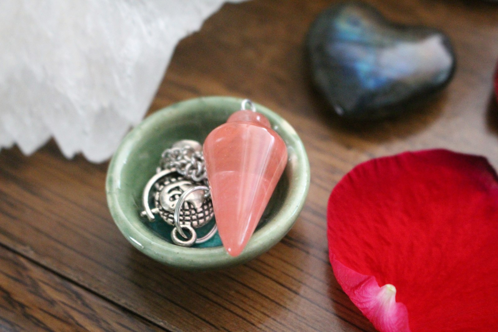 Un pendule en pierre rose posé dans une petite coupelle en céramique, à côté d'un cristal blanc, d'une pierre sombre et d'un pétale de rose