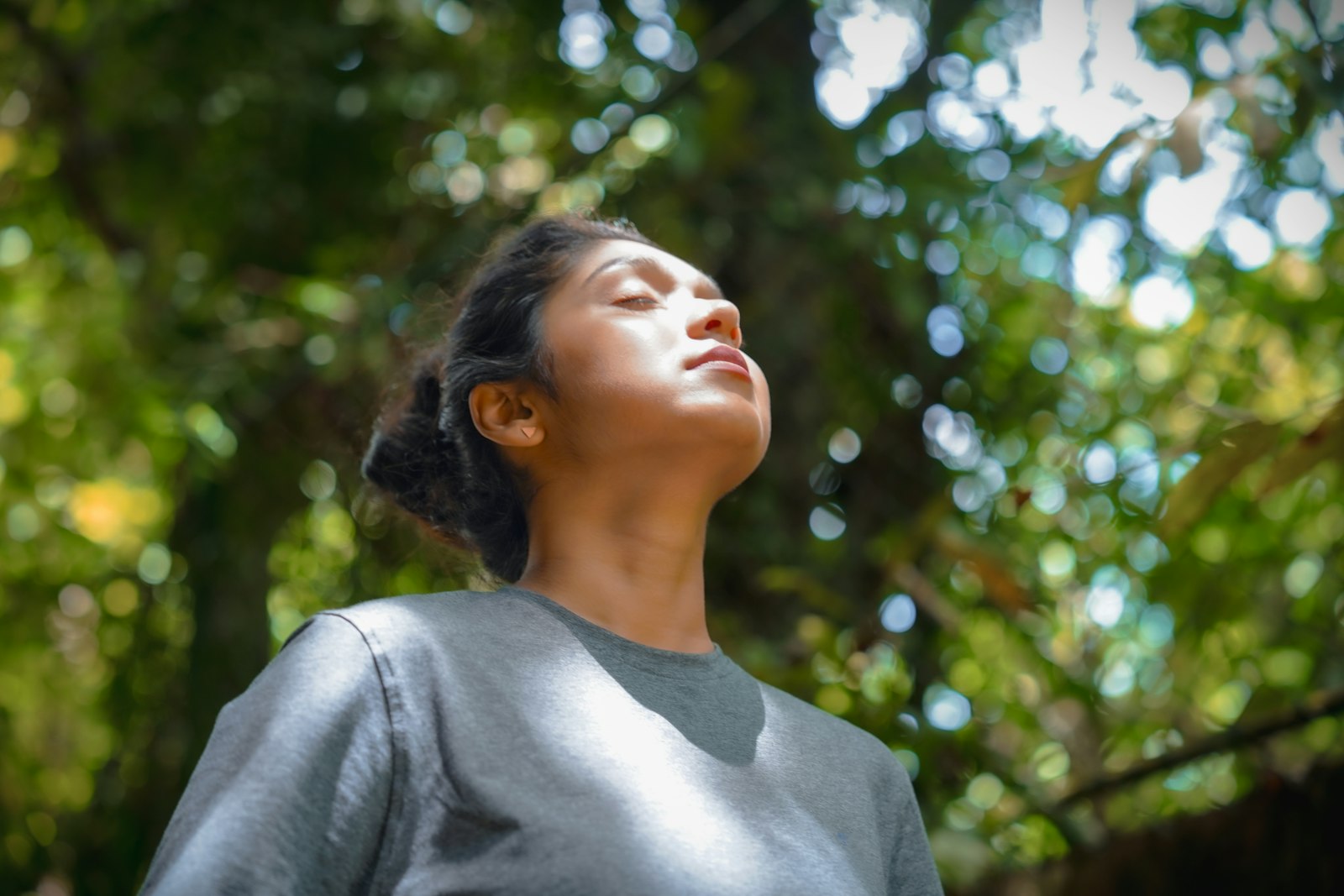 Femme les yeux fermés qui respire profondément sous les arbres, le visage tourné vers la lumière